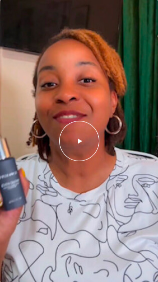 A woman with light brown locs is smiling and holding a dark blue bottle of Even Glow