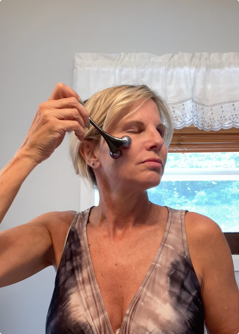 A woman with short blonde hair is using a facial roller on her face in a bathroom.