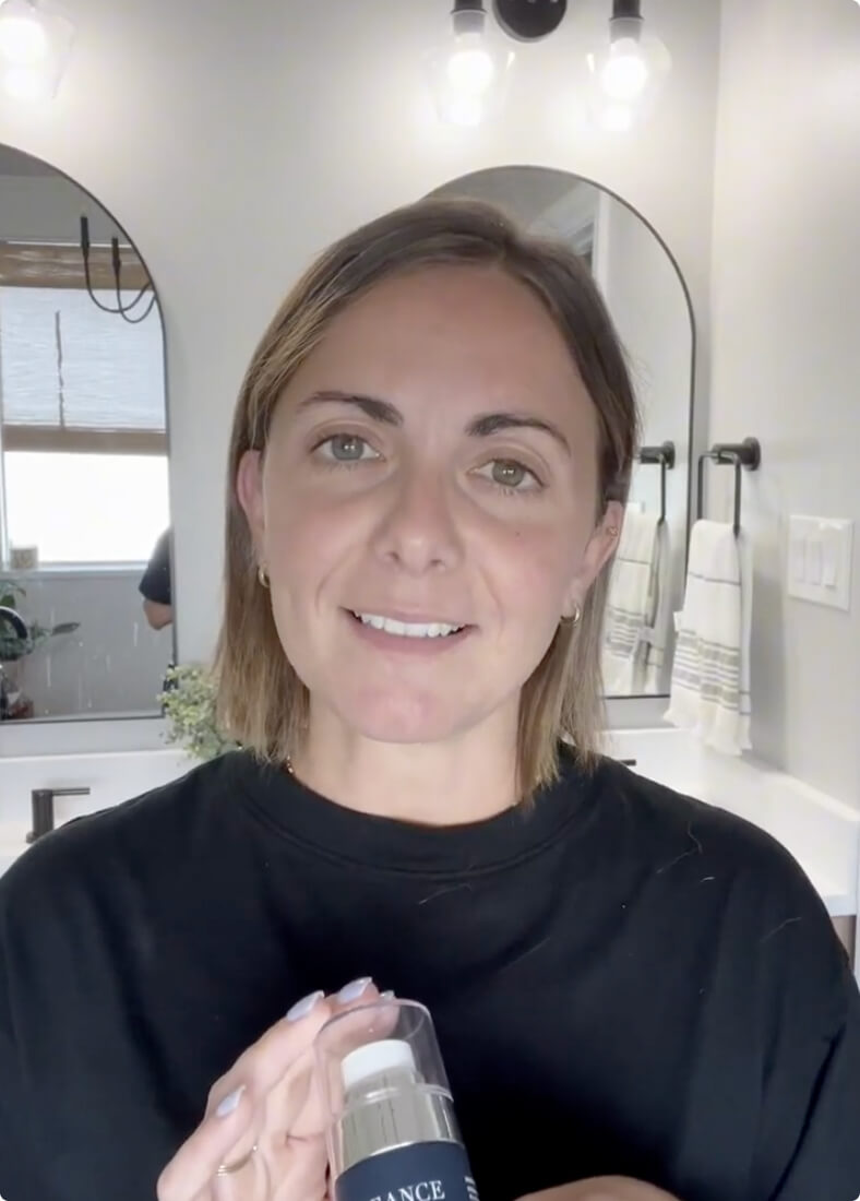 A woman with short brown hair is standing in a bathroom holding a bottle of Lift & Glow serum.