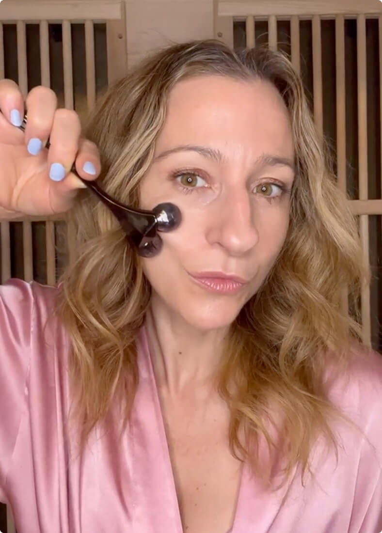 A woman with wavy blonde hair uses a Contour Massage Roller on her cheek.