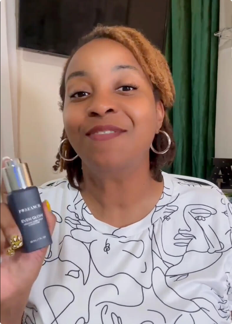 A woman with light brown locs is smiling and holding a dark blue bottle of Even Glow.