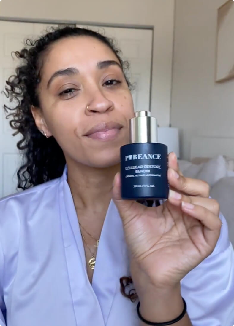 A woman with curly hair holds a dark blue bottle of Pureance Cellular Restore Serum.