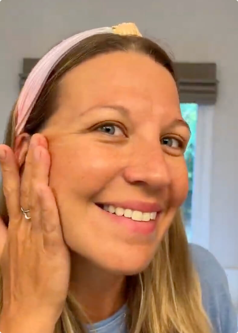 A smiling woman with long blonde hair and a pink headband touches her cheek.