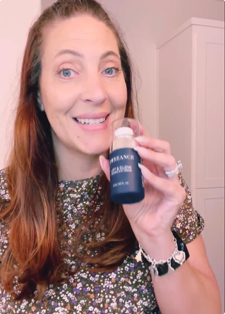 A woman with long, light brown hair is smiling and holding a bottle of Lift & Glow serum.